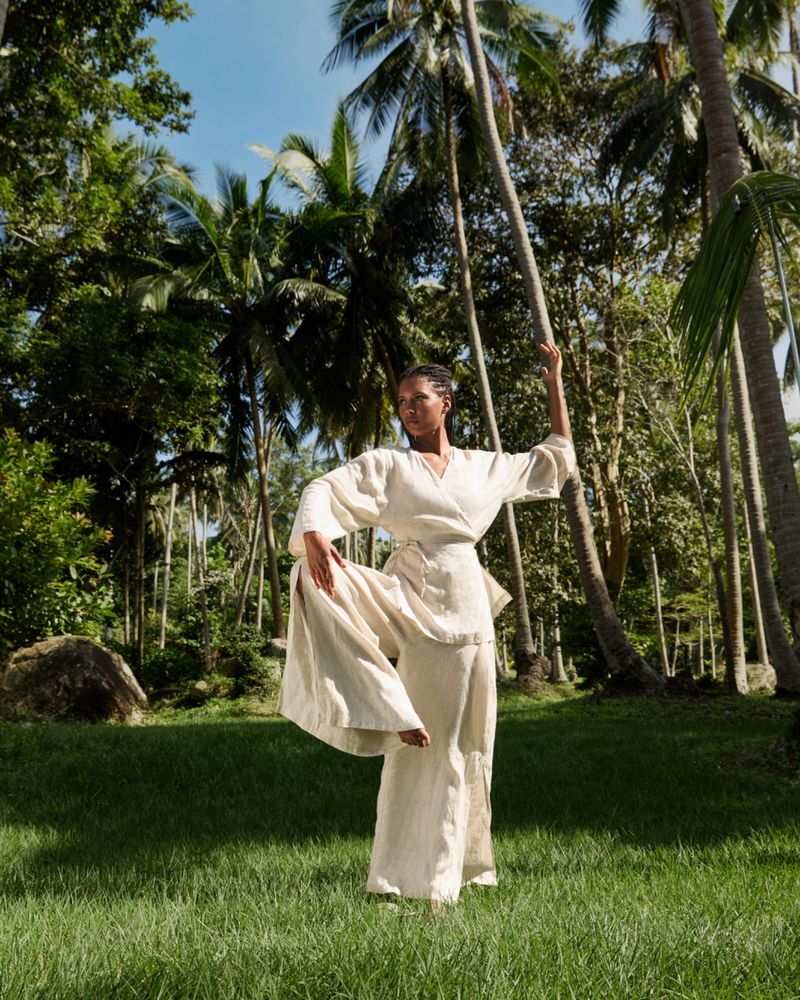 Woman practices yoga outdoors in flowing white clothing, with a leg raised. Palm trees and lush tropical plants surround the scene.