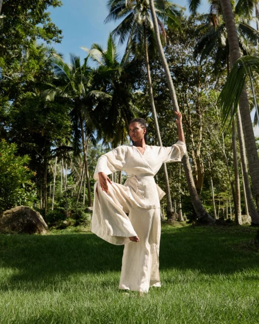 Woman practices yoga outdoors in flowing white clothing, with a leg raised. Palm trees and lush tropical plants surround the scene.