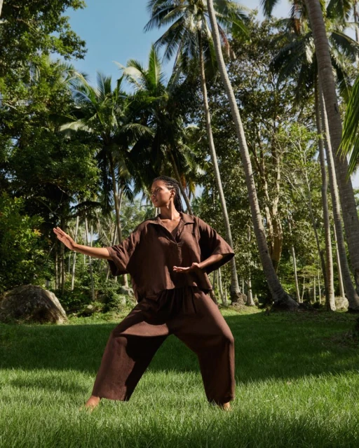 Woman in brown outfit stands in a relaxed fighting pose in a grassy field surrounded by tall palm trees and lush green foliage.