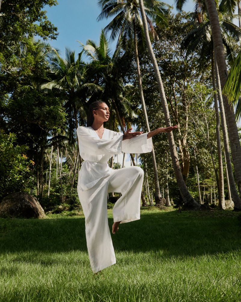 Woman in a white outfit in a yoga pose in a grassy field, surrounded by palm trees and dense tropical foliage in bright daylight.