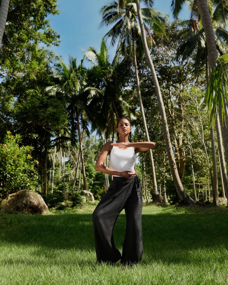 Young woman in white top and black wrap pants stands in a yoga pose on green grass, surrounded by palm trees and blue sky.