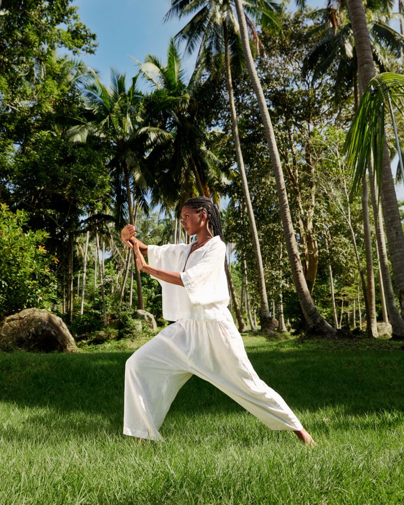 Adult female in white clothing practices martial arts in a grassy, tropical environment with palm trees and dappled sunlight outdoors.