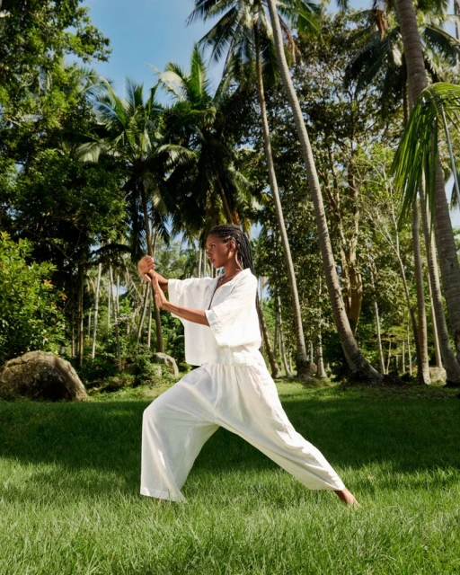 Adult female in white clothing practices martial arts in a grassy, tropical environment with palm trees and dappled sunlight outdoors.