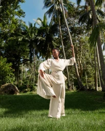 Woman wearing a beige linen wrap top and matching trousers posing gracefully amidst tall palm trees in a lush grassy clearing.