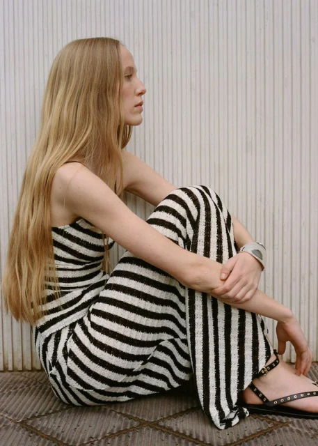 Woman with long blonde hair sitting, wearing black and white striped pants and top with black sandals against a white striped wall.