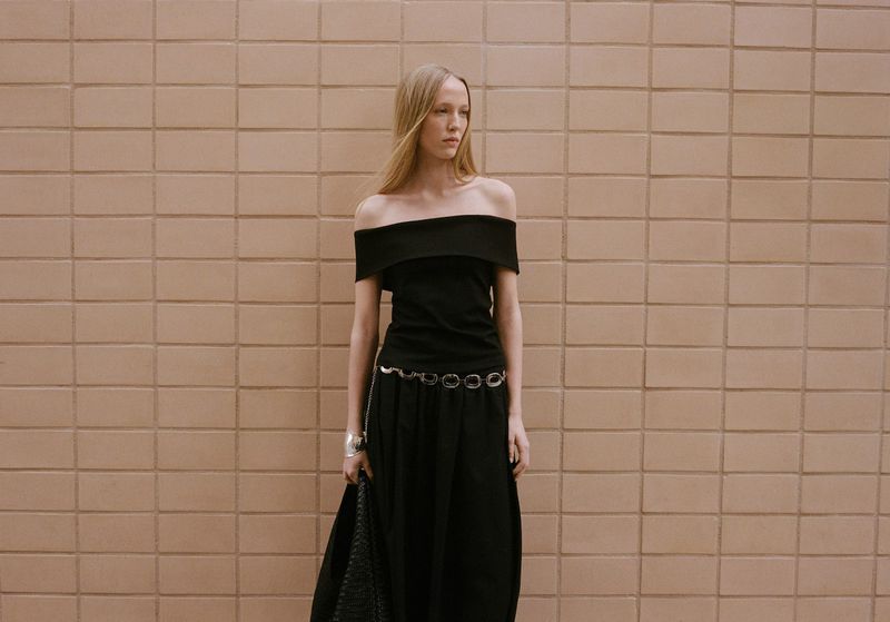 An adult woman poses in a black off-the-shoulder top and long skirt against a tan brick wall, wearing a silver cuff and belt.