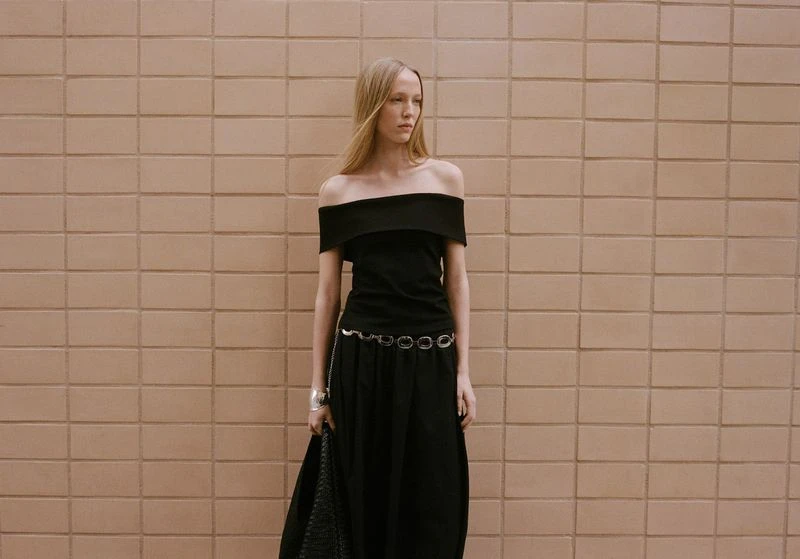 An adult woman poses in a black off-the-shoulder top and long skirt against a tan brick wall, wearing a silver cuff and belt.