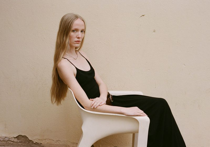 Woman with long blond hair sits in a modern chair wearing a black dress against a tan stucco wall in a fashion editorial shot.