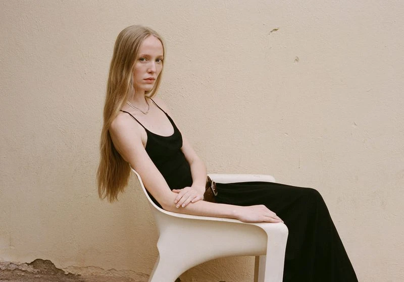 Woman with long blond hair sits in a modern chair wearing a black dress against a tan stucco wall in a fashion editorial shot.