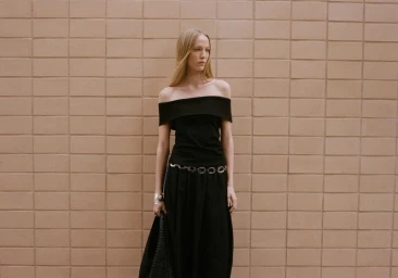 Model standing against a tiled brick wall wearing a black off-the-shoulder top and black skirt with chain belt.
