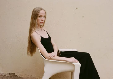 Model with long blonde hair seated in a white plastic chair wearing a black dress against a neutral textured wall.