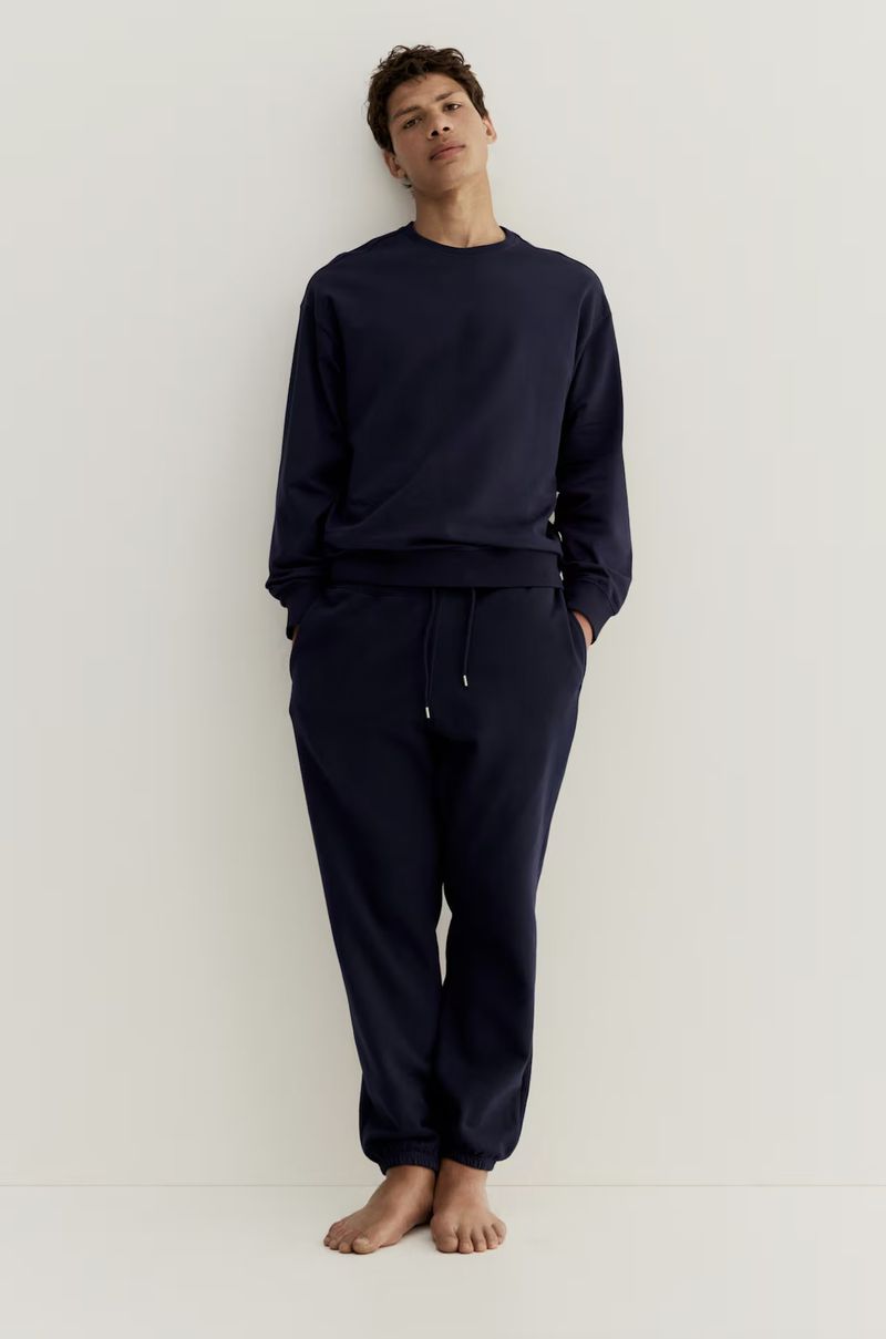 Adult male leaning against a white wall wearing a navy blue sweatshirt and sweatpants, indoors in a casual pose.