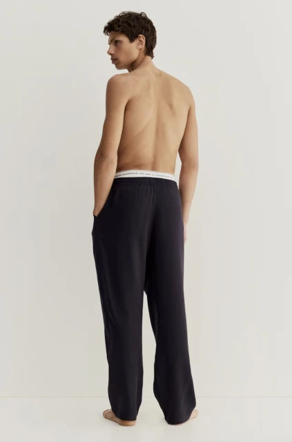 Adult male wearing dark lounge pants stands in a neutral, indoor studio setting, showcasing a relaxed and comfortable style.