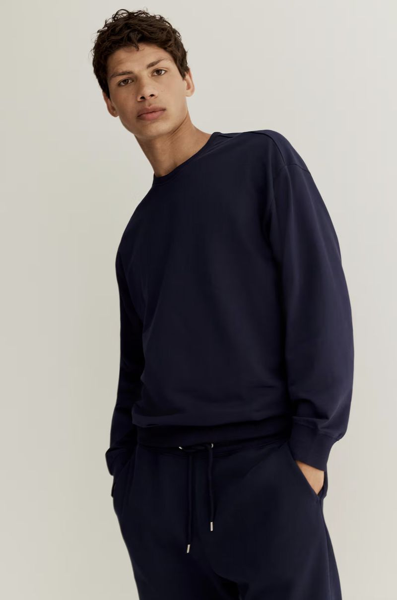 Male model wearing a navy blue sweatshirt and matching sweatpants against a plain white seamless studio background.