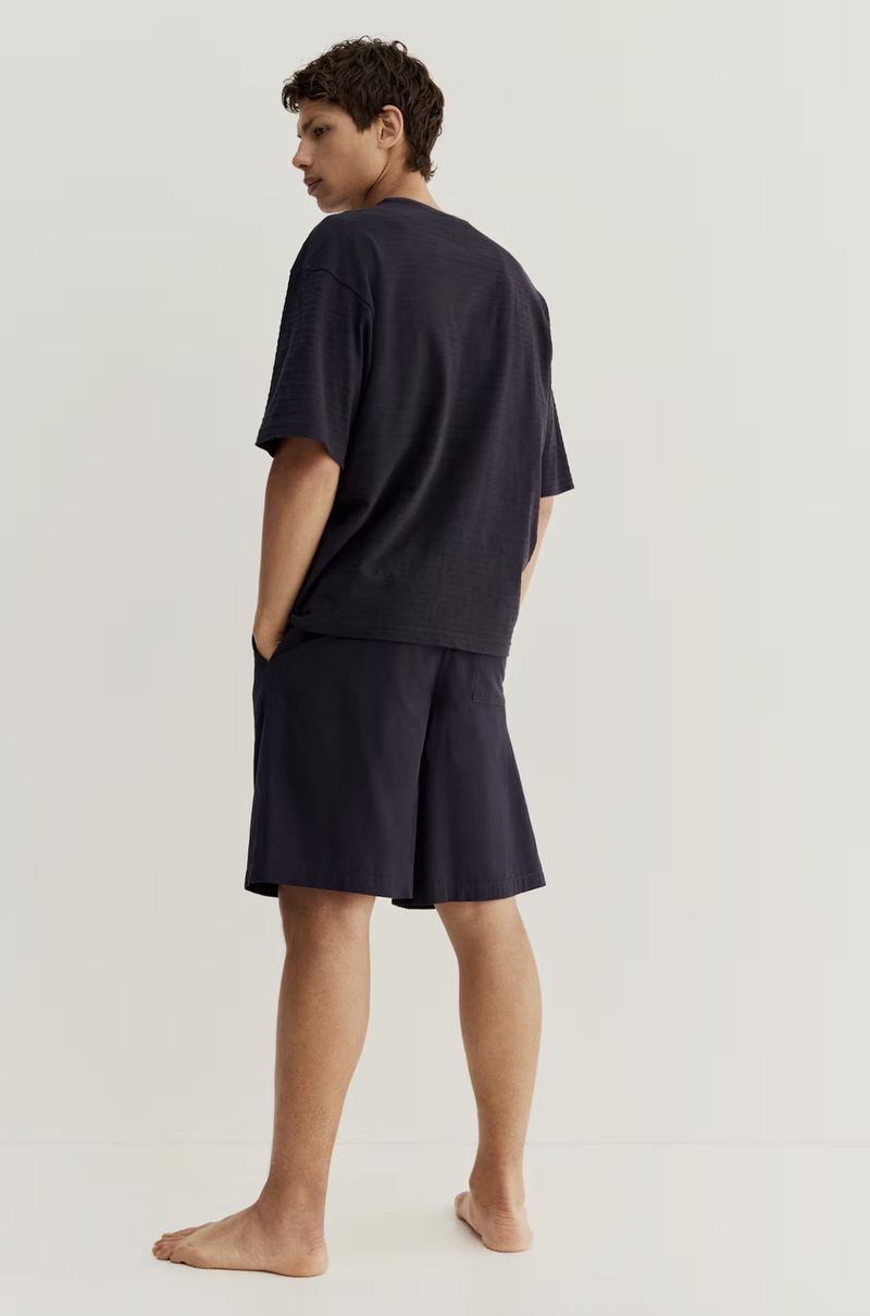 Adult male stands indoors in a relaxed pose, wearing a dark blue t-shirt and shorts set against a neutral background.