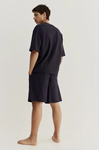 Adult male stands indoors in a relaxed pose, wearing a dark blue t-shirt and shorts set against a neutral background.