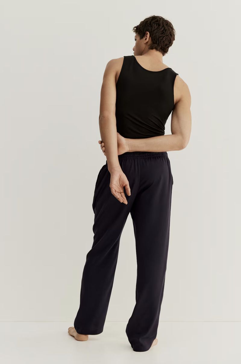 Athletic adult male with dark hair and black tank top poses in relaxed posture, back to the camera, wearing navy lounge pants in studio.