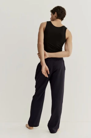 Athletic adult male with dark hair and black tank top poses in relaxed posture, back to the camera, wearing navy lounge pants in studio.
