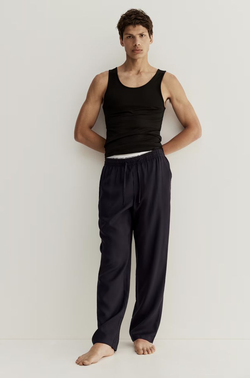 A slender man poses indoors wearing a black tank top and navy drawstring pants against a pale backdrop in soft, diffused lighting.