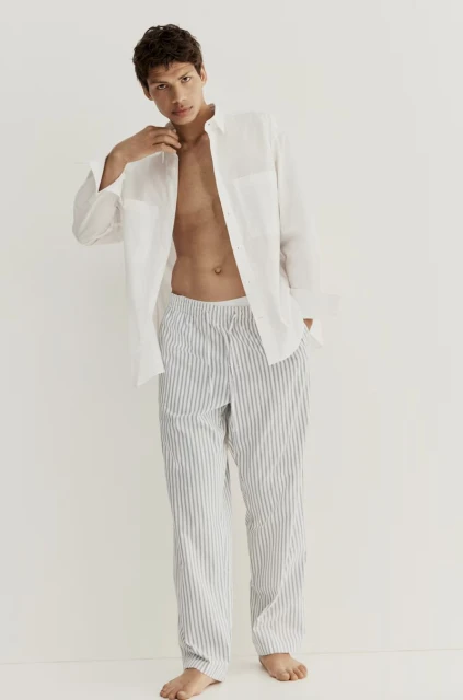 An adult man in a white button-down shirt and blue and white striped pants stands in a bright studio, posing for a fashion shot.