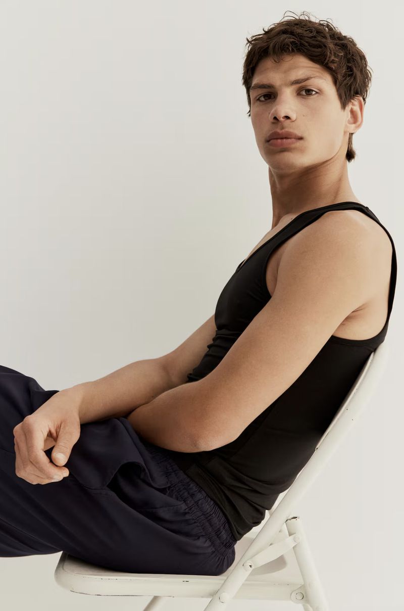 A male model wearing a black tank top and dark trousers seated on a white folding chair against a plain background.