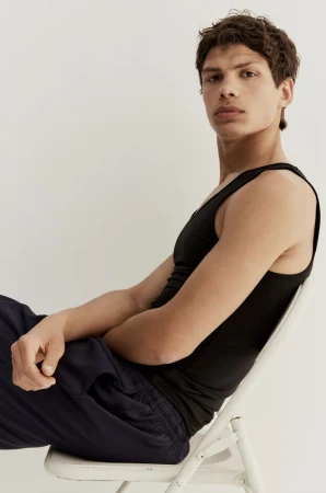 A male model wearing a black tank top and dark trousers seated on a white folding chair against a plain background.