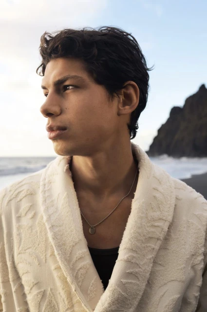 Portrait of a young adult male wearing a terry cloth robe on a beach with black sand, with a rocky cliff in the background.