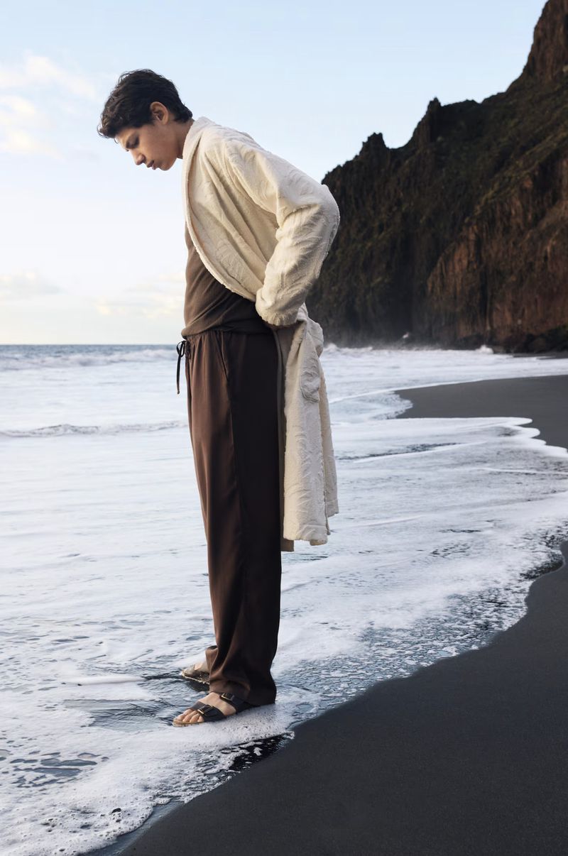 Young man in casual attire standing on a black sand beach with ocean waves and a rocky cliff in the background during daylight.