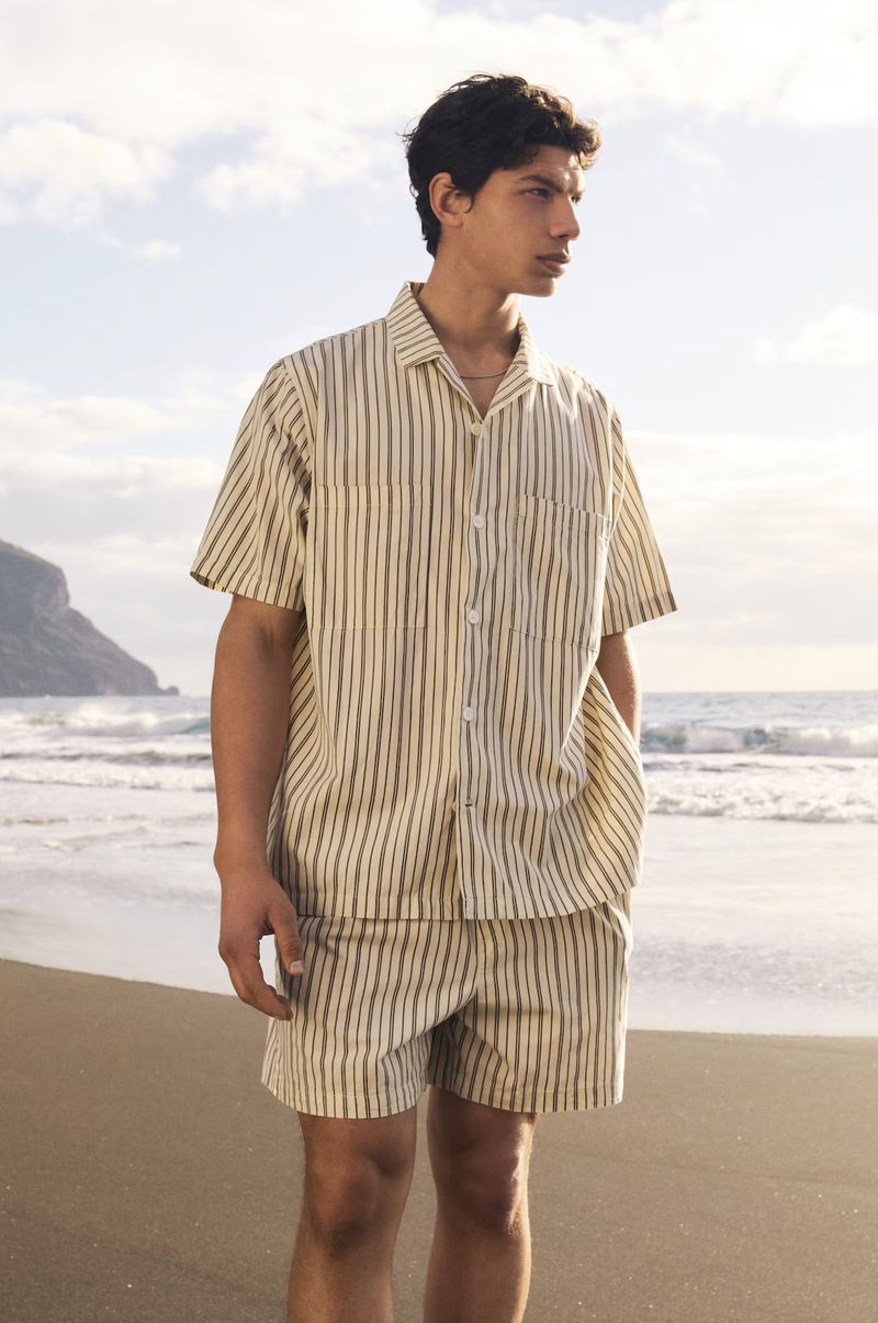 Male model wears striped shirt and shorts on a beach with ocean waves, blue sky, and distant hills. Fashion, outdoors, summer style.