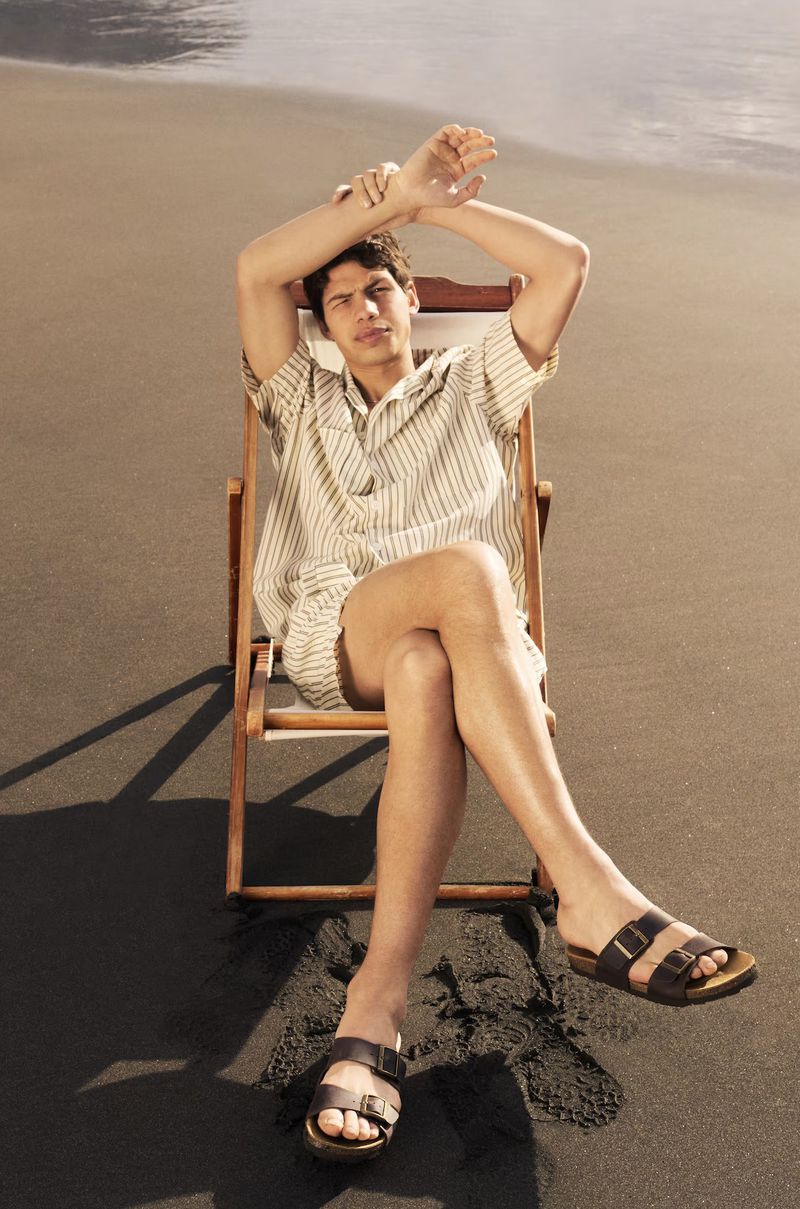 A man relaxing in a wooden deck chair wearing sandals on a black sand beach with ocean water in the background.
