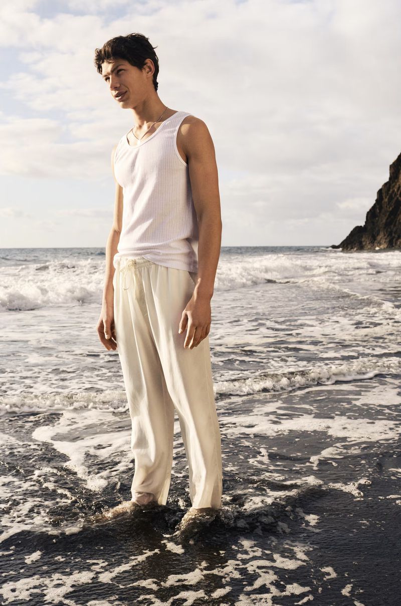 Male model wearing a white tank top and cream trousers standing on a dark sandy beach with ocean waves in the background.