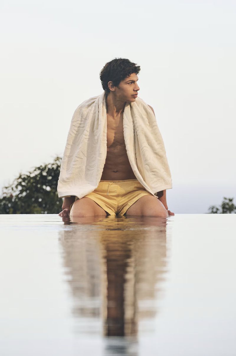 Man sitting on edge of infinity pool wearing shorts and a towel, looking out over the horizon, with reflection visible in the water.