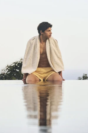 Man sitting on edge of infinity pool wearing shorts and a towel, looking out over the horizon, with reflection visible in the water.