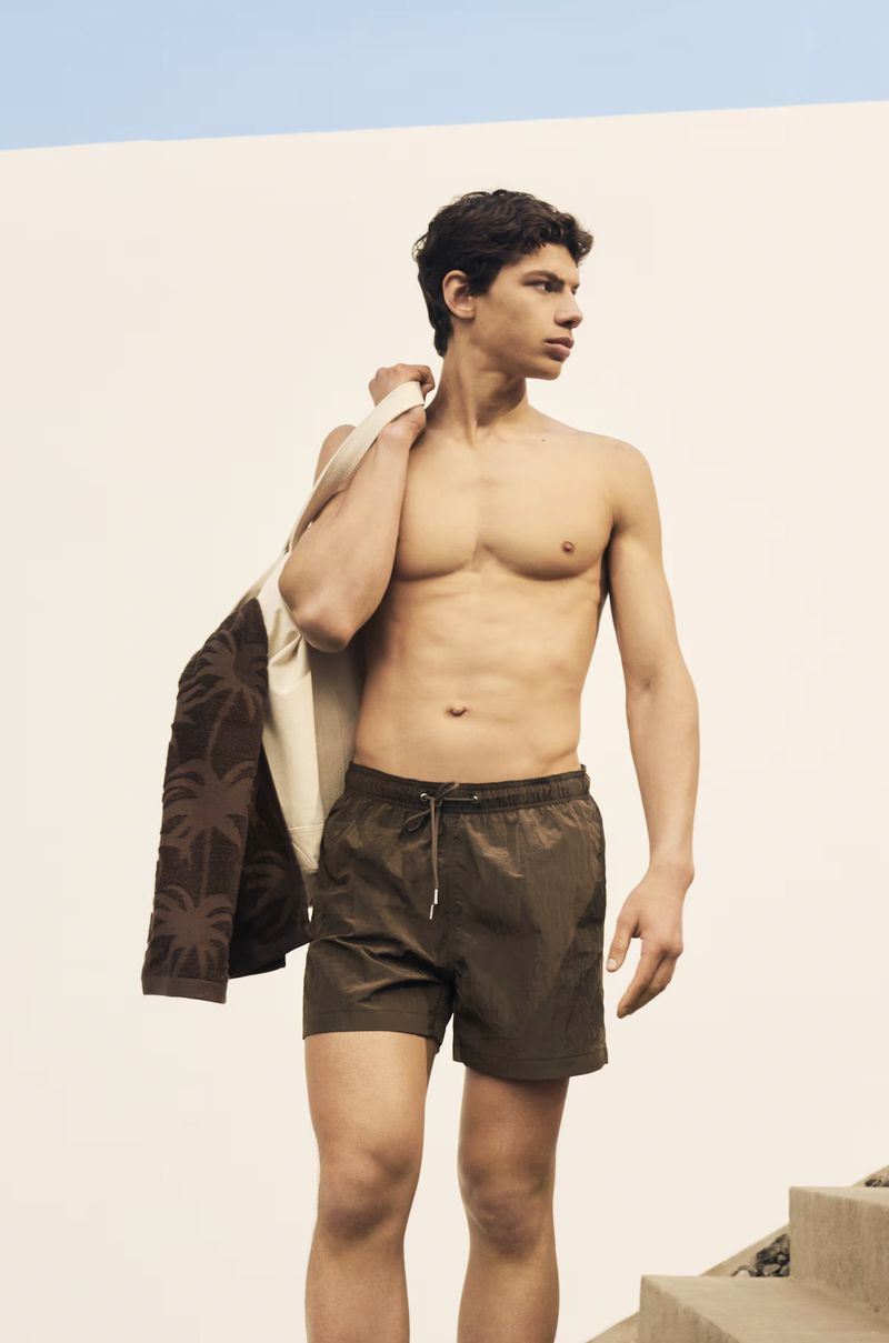 A tan-skinned adult male in olive swim trunks stands outside against a white wall and blue sky while holding a beach bag.