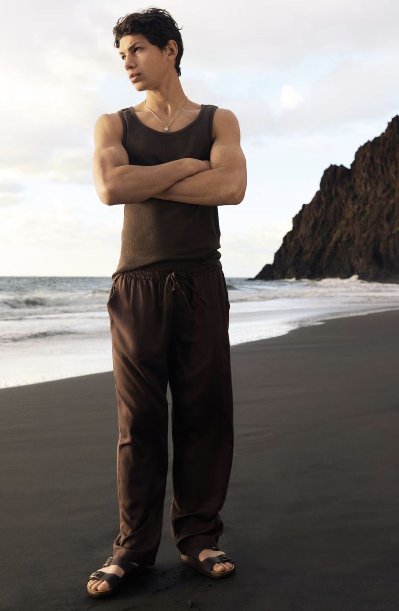 Young man stands with arms crossed on a black sand beach near the ocean with a dark rock formation in the background.