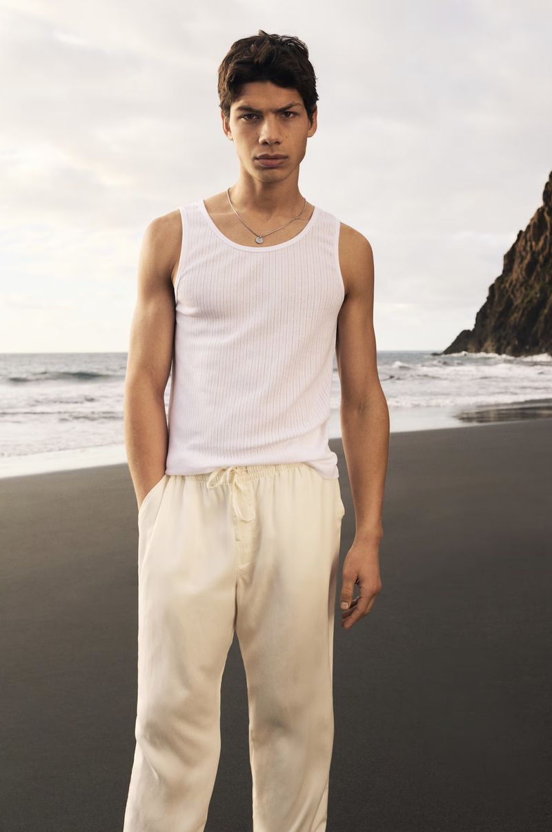 Portrait of a young adult male model in a tank top and pants on a beach with cliffs in the background, under an overcast sky.