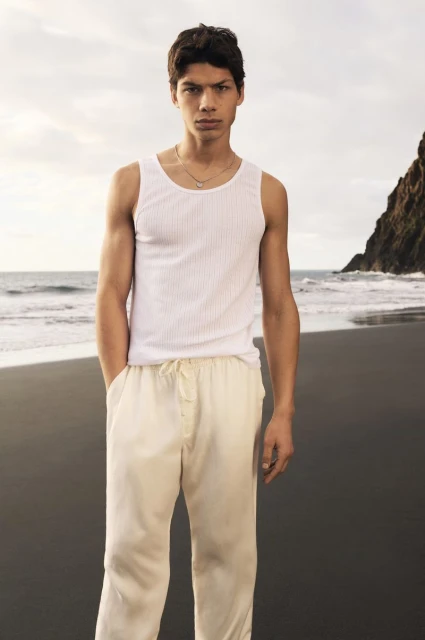 Portrait of a young adult male model in a tank top and pants on a beach with cliffs in the background, under an overcast sky.