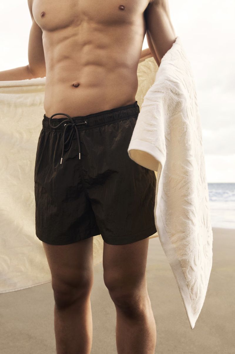 Man wearing dark swim trunks holding a white towel against a sandy beach and ocean backdrop