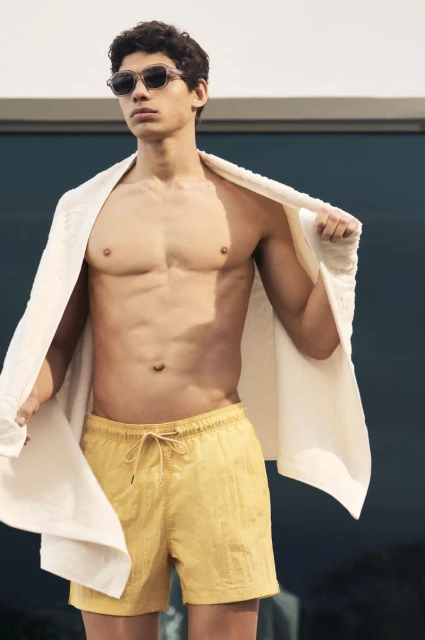 Man with sunglasses, shirtless, draped towel, and yellow shorts stands outdoors on a sunny day in medium shot for fashion concept.
