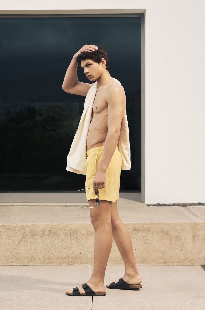 Male model in yellow shorts and sandals poses outdoors with a towel over his shoulder in front of a modern building and steps.