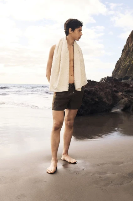 Barefoot adult man stands on beach wearing dark shorts and white towel, looking out at ocean and rock formations on overcast day.