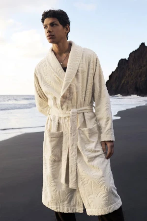 Man in a textured cream-colored bathrobe stands on a black sand beach with a rocky outcrop and ocean waves in the background.