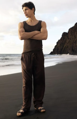 Model standing on a black sand beach wearing a brown tank top and trousers against an ocean background.