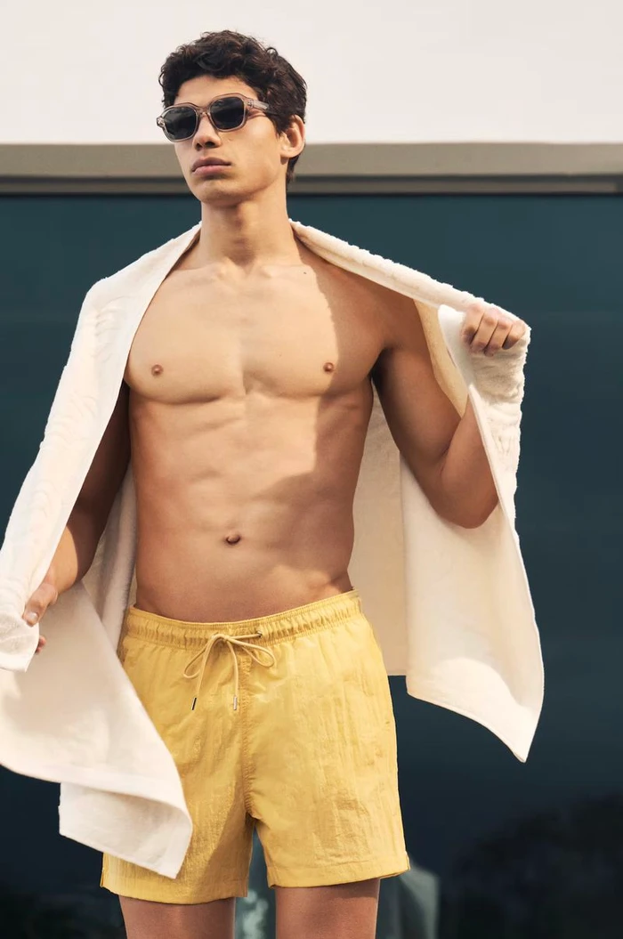 Shirtless male model wearing yellow swim trunks and sunglasses with a white towel draped over his shoulders outdoors.