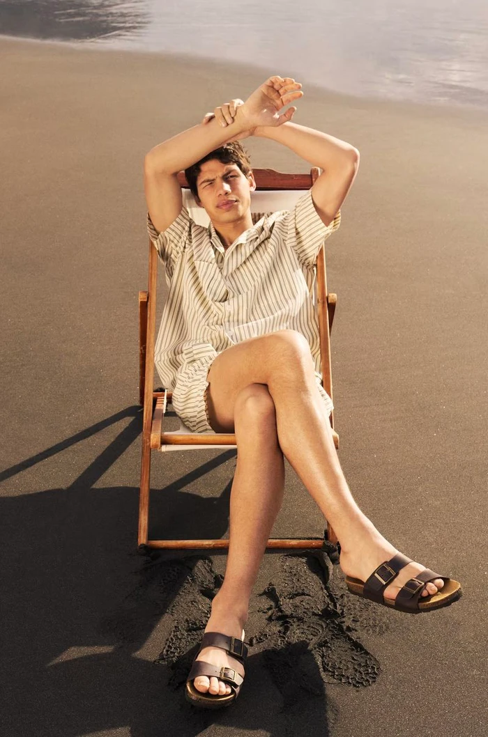 Male model seated on a wooden deck chair on dark sand wearing a striped shirt and shorts with sandals.