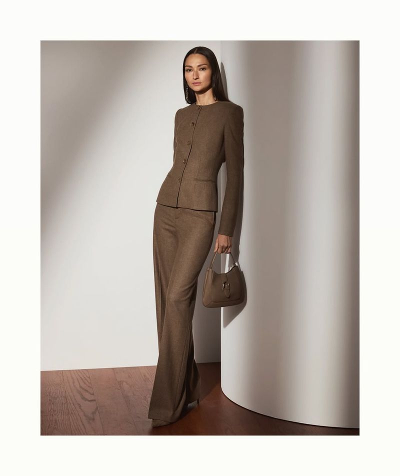 A woman in a brown suit and heels poses in front of a white wall with a matching purse, illuminated by soft studio lighting.