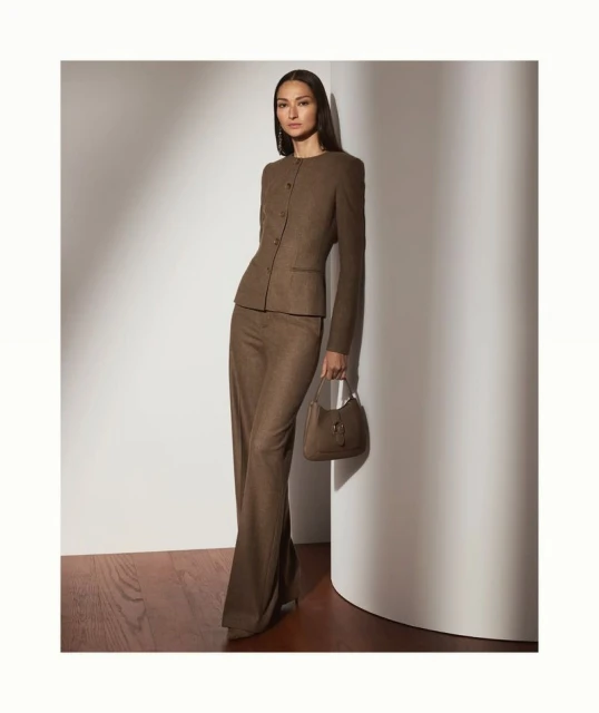 A woman in a brown suit and heels poses in front of a white wall with a matching purse, illuminated by soft studio lighting.