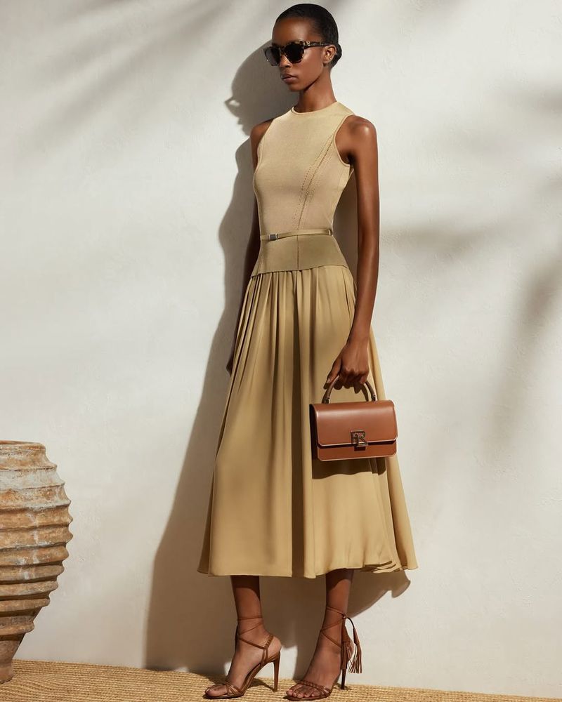 A slender fashion model in a tan sleeveless dress with a matching handbag and strappy heels poses in front of a textured wall.