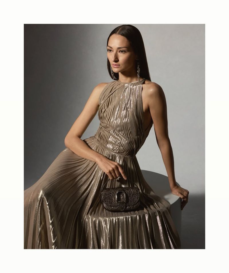 Bruna Tenorio seated in a metallic pleated gown holding a handbag against a grey backdrop for Ralph Lauren Pre-Spring 2025