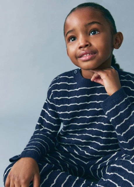 A young child poses indoors wearing a navy striped knit outfit, looking up with a neutral facial expression, captured in a studio setting.
